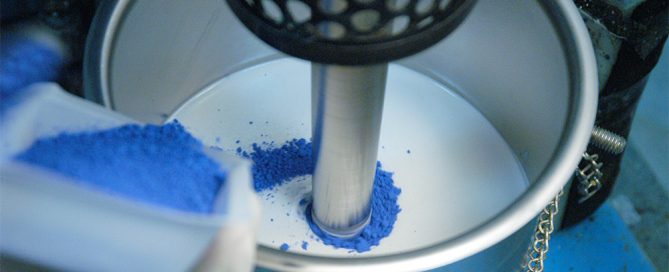 Vibrant blue pigment powder being stirred in a white bucket with additional powder on the surface.