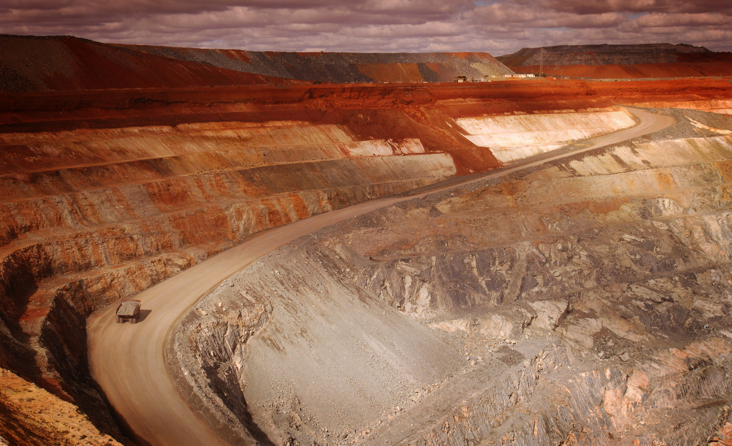 A wide angle elevated view of an open cut mine.  An excavator scoops up dirt and ore and is about to empty it into the rear tray of the haulage truck.