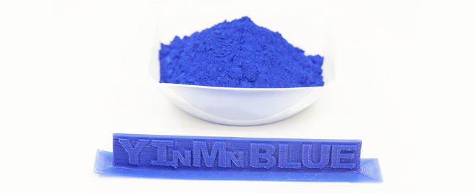 White bowl of vivid YInMn Blue powder pigment with a matching blue pigmented sample stick below.