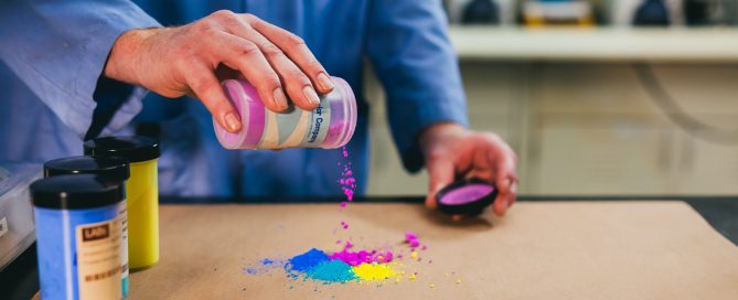 Glass jars of vibrant liquid pigments in yellow, blue, and green with small measuring tools suggesting lab formulation work.