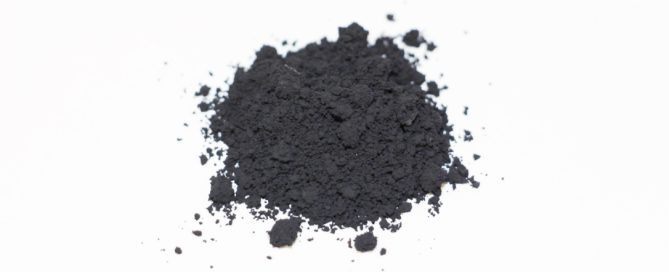 Pile of dark charcoal or black pigment powder against a white background with scattered granules and subtle shadowing.