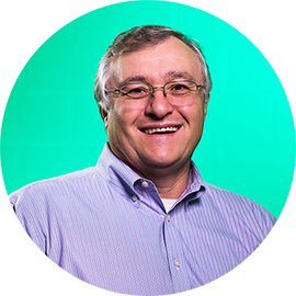 Professional headshot of a man with gray hair and glasses wearing a lavender shirt against a turquoise-green circular background.