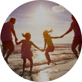 Family of four playing in shallow ocean water at beach sunset, silhouetted against warm golden sky.