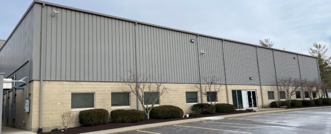 Exterior of an industrial warehouse with corrugated metal siding, tan brick foundation, and landscaping on an overcast day.